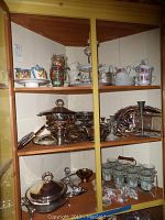 Shelf showing an assortment of plates, silver-tone serving dishes, decorative cups, and a multi-tier serving platter stand.