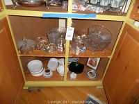 Shelf holding assorted cut glass mugs, bowls, dishes, some white ceramic dishes on lower shelf.