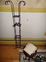 Tall metal wall hanger standing on floor against wallpapered wall, with paper napkins on small stool in front