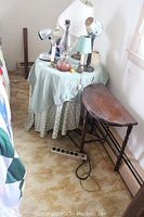 Photo showing two side tables; one covered with cloth and various lamps, the other a wooden demi lune side table with claw feet and a curved half-moon top, placed against the wall.