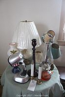 Full view of assorted lamps displayed on a fabric-covered table including oil lamps, candle sticks, lamp shades, and ceramic lamp bases.