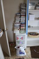 Tall stack of assorted greeting cards and boxes on top of white plastic storage drawers, situated near a doorway with a bag on the floor.