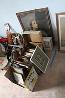 Cardboard boxes filled with assorted framed prints and paintings of various sizes, some leaning against the wall, including a large portrait painting and smaller framed art.