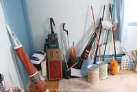 Wide shot showing multiple cleaning tools including two Hoover vacuums, various brooms, mops, dust pans, dusters, and four decorative buckets arranged against a wall in a corner of a room.