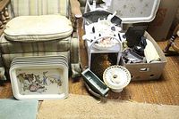 Wide view of the lot with decorative TV trays stacked, religious figurines, vintage book holders, ceramic planters, and a box of miscellaneous decor items arranged on carpet next to an armchair.