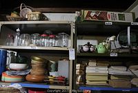 Metal shelves holding assorted glass jars, wooden bowls, plastic trays, colorful ceramic plates and stacked paper boxes below.