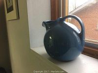 Side view of large blue ceramic pitcher on a windowsill showing round shape and handle.