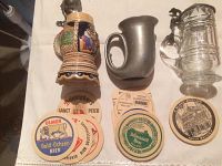 Three beer steins including one ceramic, one pewter marked USA, and one glass with domed lid, plus assortment of beer coasters.