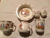 Top-down photo showing Bunnykins cereal bowl with decor of rabbits on rim and a Bunnykins scene inside. Also visible are various Bunnykins cups: one two handled cup, two cake cups, and two drinking cups with Bunnykins character illustrations.