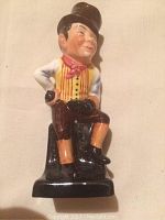 Porcelain figurine showing a seated man in a yellow and red striped vest, brown pants, black boots and brown top hat.