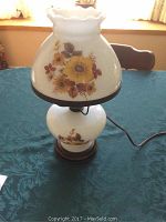 Photo of a vintage glass dual light table lamp with floral artwork in yellow and brown colors, showing the entire lamp including base, shade, and power cord on a blue tablecloth.