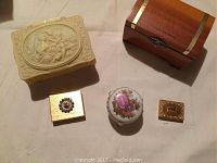 Five trinket boxes: cream-colored ornate box with cherub relief, footed Limoges floral box, wooden chest box with brass trim, two small gold-toned decorative boxes.