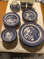 Set of dinner plates, small and large bowls, cups, and saucers arranged on a cloth-covered surface showing Blue Willow pattern.