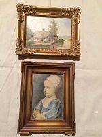 Photo showing both framed pictures laid flat, one vertical portrait of a child and one horizontal cottage landscape with damaged frame corner.