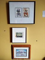 Full view of three framed artworks hung on a yellow wall: two bird-themed prints by Zhao Ziping framed together, small Manhattan skyline print and a signed print of seals by Mabel Nigiyok.