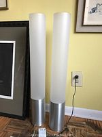 Two tall modern floor lamps standing side by side with cylindrical metal bases and tall frosted glass shades.