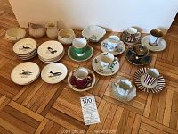 Assorted espresso cups and saucers along with several small plates and small vases or containers arranged on wood parquet floor
