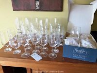 Large collection of wine glasses and stemless glasses arranged on a wooden buffet top. Includes box for Marquis Vintage glasses and several loose glasses.