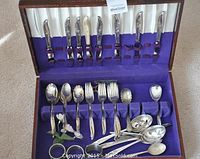 Full view of the entire silverware set inside purple lined case with knives in upper lid and spoons, forks and utensils in lower section.