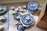 Full set of Johnson Bros Willow Blue china laid out on carpet, including plates stacked on stand, bowls, cups, and sugar bowl.