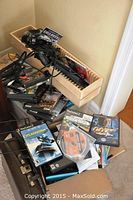 Two boxes visible with assorted media items: VHS tapes, CDs, and PlayStation 2 game discs with cases, stacked and stored in cardboard and wooden boxes.