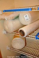 Three large rolls of bubble wrap, one roll of clear plastic wrap, multiple sheets of foam padding stored on white wire shelving.