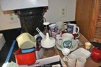 Overview of miscellaneous mugs, bowls, and kitchenware including boxed Tribest blender and metal bread pans.