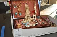 Photo showing a brown jewelry box open with assorted necklaces including pearl and turquoise, clip-on earrings hanging from hooks, and rings in slots.