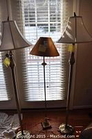 Three floor lamps: two tall matching lamps and one smaller lamp placed in front of window with horizontal blinds, showing full length and shades.