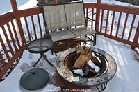 Full lot on snowy deck: glider bench, fire pit with logs, side table, umbrella base.