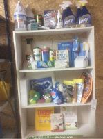 Front view of full 4-shelf white unit showing cleaning chemicals on top shelf, cleaning cloths and sprays on second shelf, assorted cleaning pads and sprays on third shelf, and stain removal and home remedy books on bottom shelf.