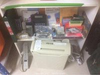Photo shows a beige paper shredder, calculators, books, power bar, and other office supplies on a shelf and floor.