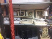 Shelf containing three framed fruit oil paintings, white drapes, metal rods, a small lamp with cream shade, and other household items.