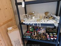 Photo of black shelving unit with assorted clear acrylic, silver and gold-toned ornaments and garlands on top shelf; brass stained glass candle holders, colorful balls, and Christmas figurines on lower shelf.