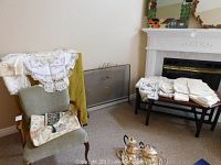 Overall view of green upholstered wood frame armchair with assorted linens draped over and around it, with fireplace and bench in background holding folded linens