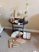 Wide shot of dark wooden side table with magazine rack, oil lamps on top, hand painted Polish eggs in a basket, Inuit bear statues, hand embroidered floral picture on easel, floral ceramic tray, and assortment of bamboo and raffia handheld fans on floor in front plus glass drip protectors in basket.