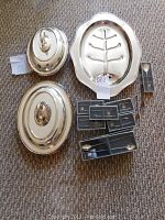 Overview photo showing two covered dishes with lids, fish skeleton tray, sets of small forks and spoons on carpet.