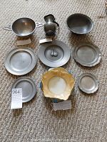Overview of all items showing nine total pieces: five pewter plates (one industrially marked), double-handled porringer, syrup pitcher, pewter bowl, and ornamental porcelain bowl.
