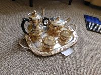 Full view of the complete silverplate tea service on the decorative tray placed on carpeted floor showing coffee pot, teapot, sugar bowl, creamer, and tray with black handles on pots.
