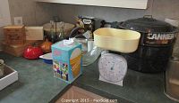 Overview of kitchen lot showing canner, kitchen scale with glass bowl, wooden boxes, red shakers, boxed ice cream machine, and other mixing bowls and utensils on kitchen counter.