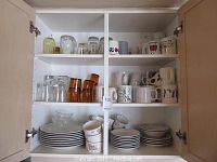 Inside kitchen cabinet showing a mix of clear glass mugs, amber glasses, ceramic mugs with different prints, stacked plates and bowls on lower shelves.