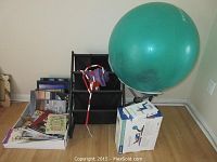 Two boxes of books on the floor next to a black tiered book rack, a green yoga ball on its box, resistance bands, and a skipping rope.