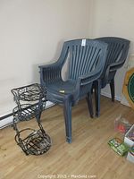 Four black plastic patio chairs stacked and black wrought metal plant stand grouped together on wood floor next to wall baseboard heater.