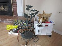 Wide shot of lot showing large metal flower-themed candle holder, gardening tools, screwdriver set, wind chimes, plastic bin with supplies, and other household items on wood floor.