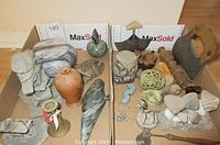 Photo showing a variety of soapstone and stone art pieces including ulu knives, frogs, carved shapes, and lidded rock container grouped in a box.