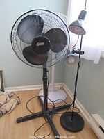 Photo of two floor standing fans (black 'Super' fan and white 'Honeywell' fan) and a 2-light floor lamp in a room corner.