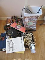 Overview of the lot with pencil crayons in different holders, black craft lamp, white plastic storage bin with assorted cords, ribbons, brushes, papers, and some crafting tools.