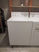 Front view showing entire white washing machine next to a dryer in a basement or utility room setting.
