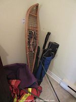 Photo showing vintage wooden snowshoes leaning against wall next to a walking stick, ski poles in blue bag, orange tent bag, folded chair and orange life jacket.