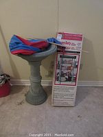 Lot showing new boxed 5-shelf steel storage rack, grey plastic bird bath with kite on top, all indoors against beige wall.